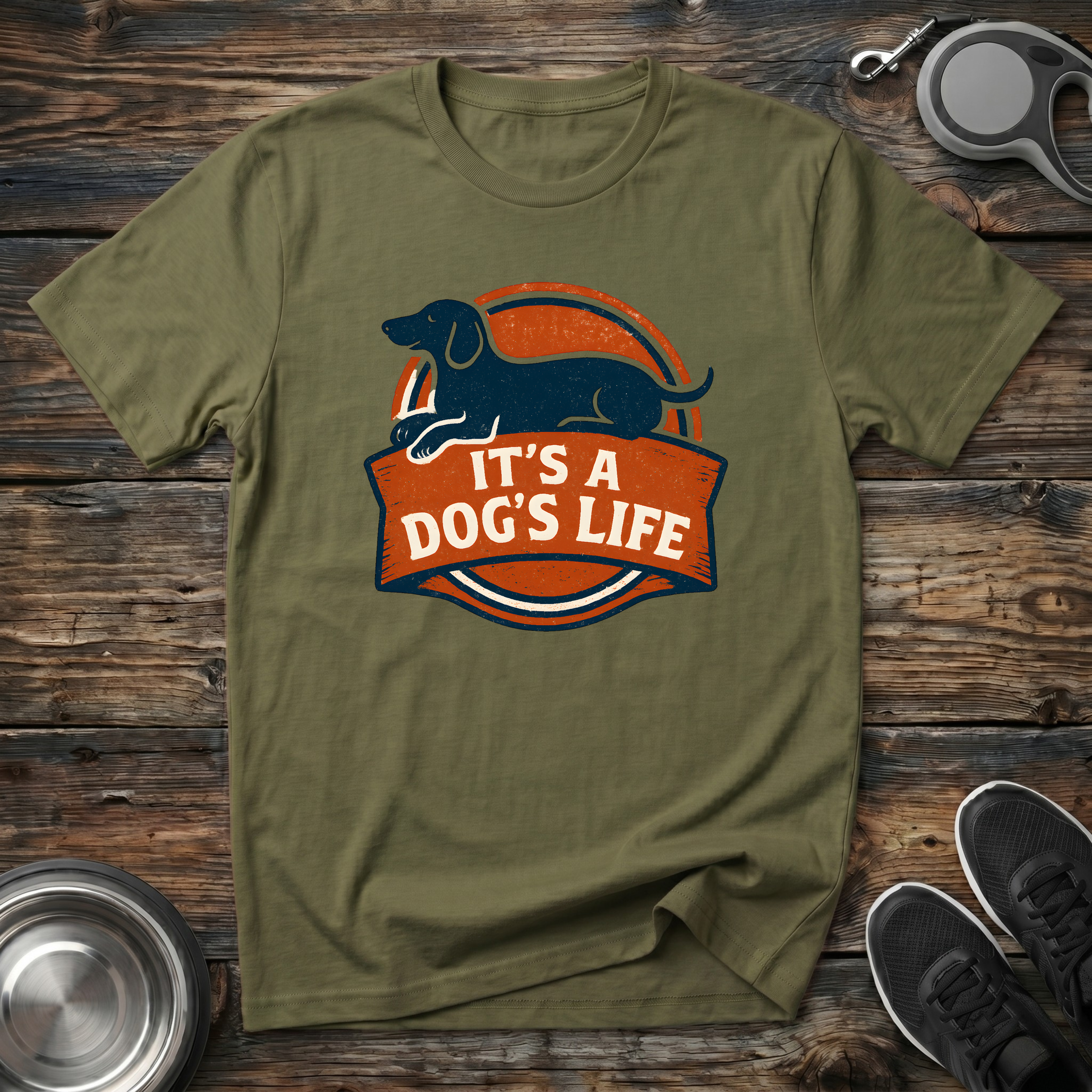 Olive green unisex t-shirt with navy Dachshund silhouette on a bold burnt-orange retro badge graphic and "It's A Dog's Life" banner lettering — vintage wiener dog lover gift tee