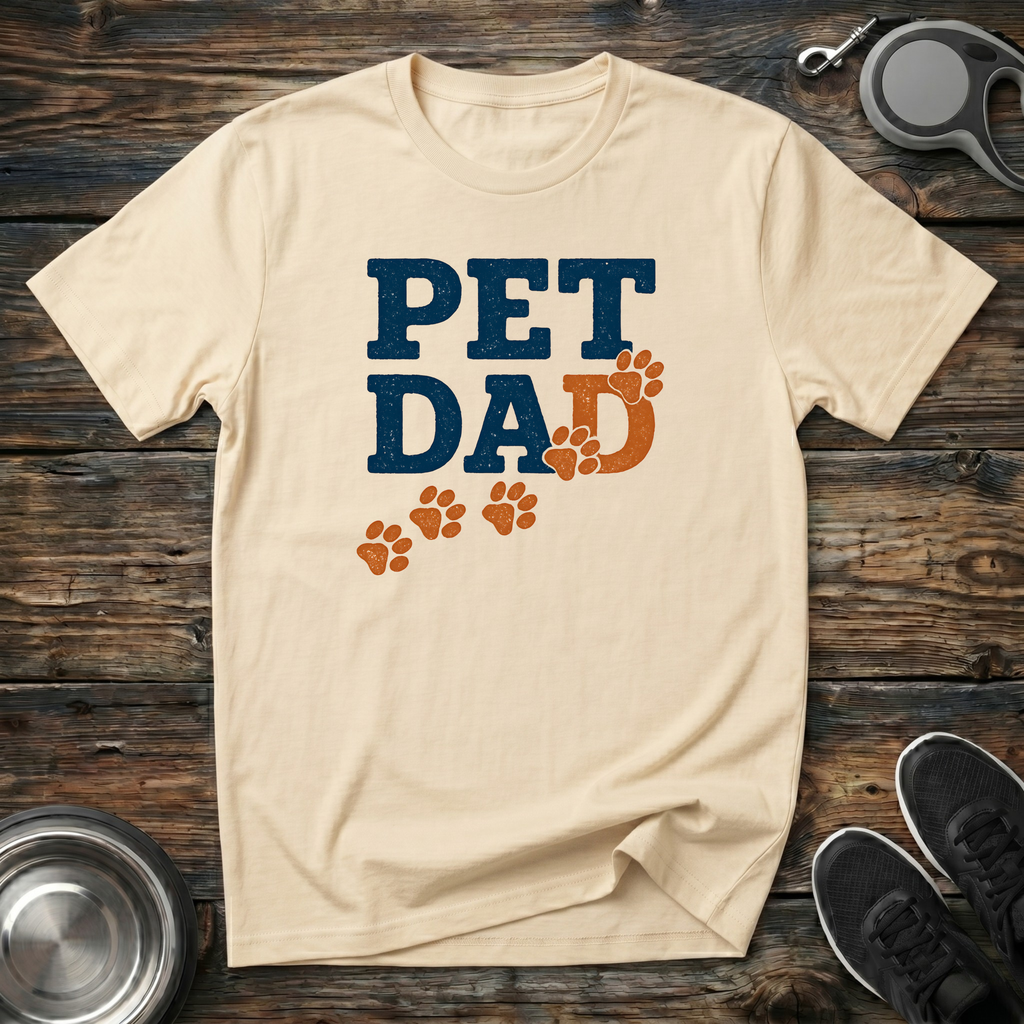 Cream unisex t-shirt with distressed navy blue "Pet Dad" block lettering and burnt-orange paw print trail graphic — funny dog dad gift tee for pet parents
