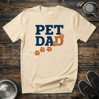 Cream unisex t-shirt with distressed navy blue "Pet Dad" block lettering and burnt-orange paw print trail graphic — funny dog dad gift tee for pet parents