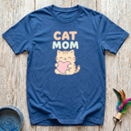 Denim blue unisex t-shirt with kawaii orange tabby cat hugging a pink heart illustration and soft peach and mint "Cat Mom" rounded lettering — cute cat lover gift tee for cat mamas